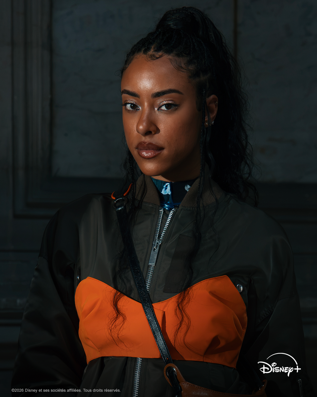Une femme aux longs cheveux bouclés, vêtue d’une veste orange et noire, se tient debout à l’intérieur, l’air impassible. Le logo Disney+ est visible dans un coin de l’image, laissant entendre qu’elle pourrait figurer dans « Un Automne Pour Te Pardonner », le dernier roman phénomène qui captive le public.