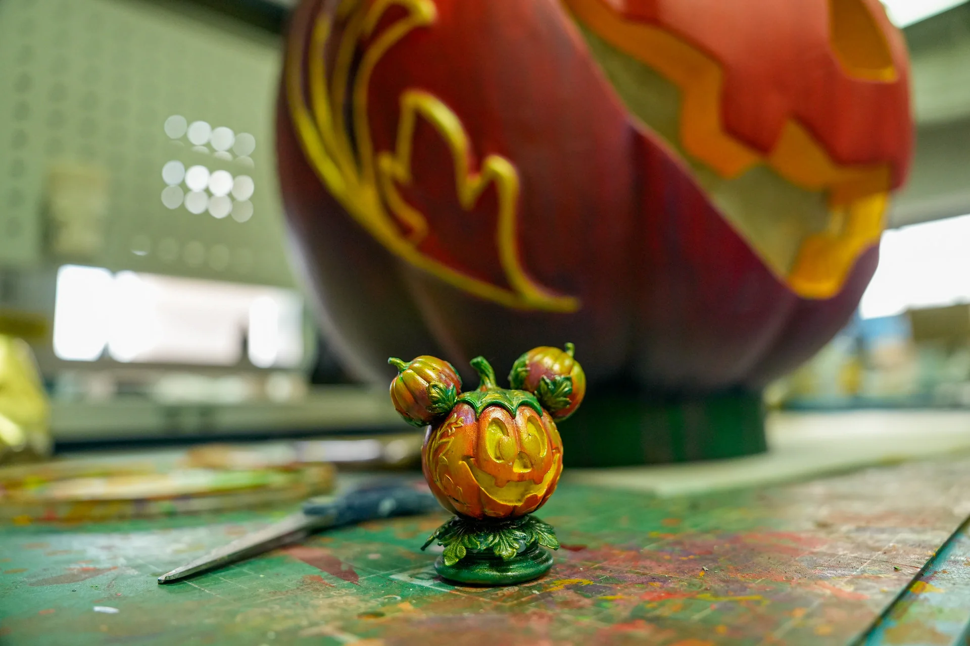 Une petite figurine en forme de citrouille, au visage souriant et coiffée de deux citrouilles, trône sur une table peinte ; une grande citrouille sculptée se détache sur l'arrière-plan flou, illustrant l'ambiance festive du Festival d'Halloween à Disneyland Paris.