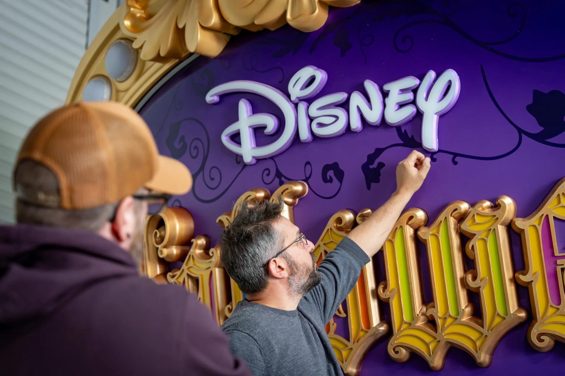 Deux personnes travaillent sur une enseigne décorative arborant le logo Disney en lettres blanches sur fond violet, agrémentée de détails dorés, en vue du féérique Festival d'Halloween à Disneyland Paris.