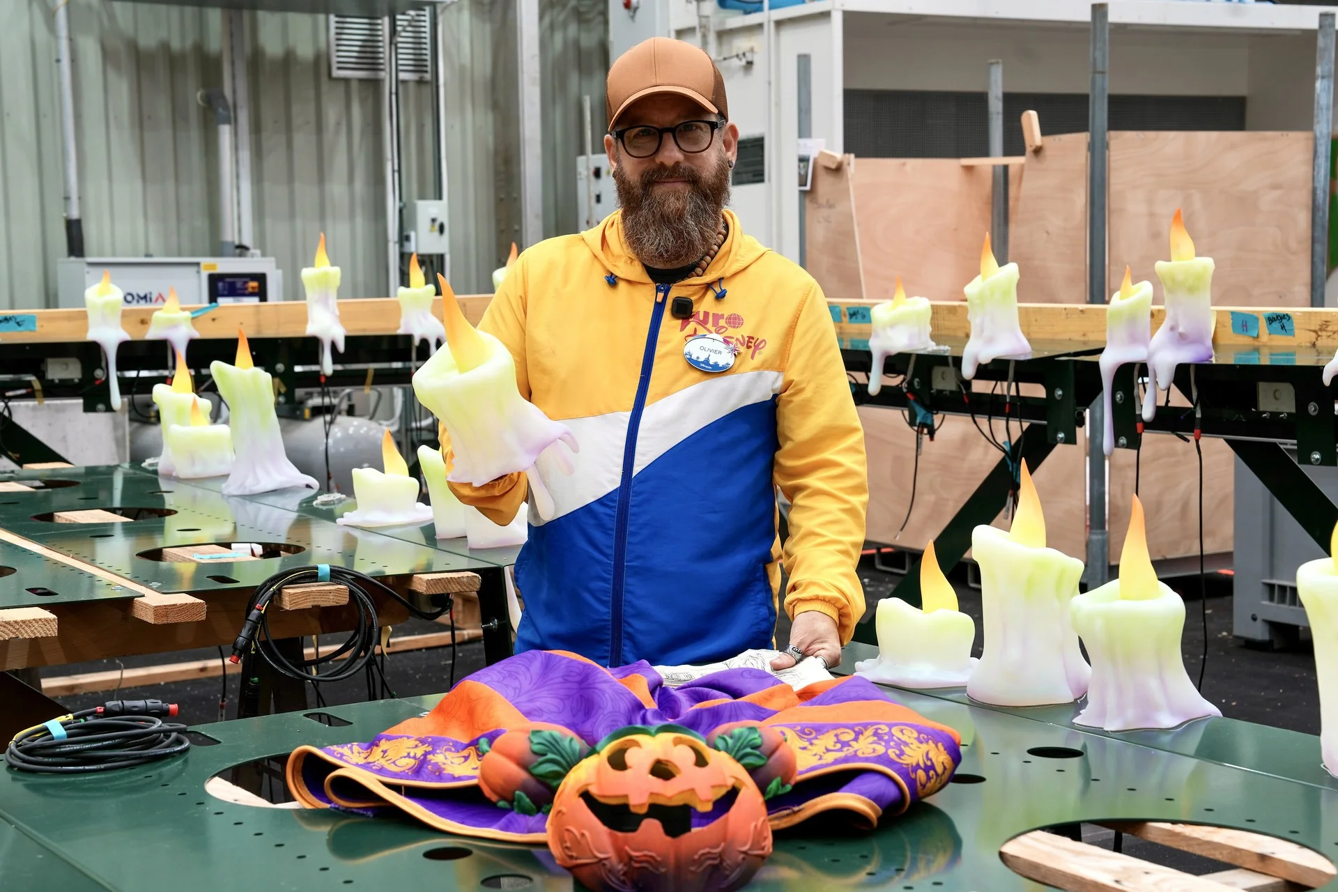 Dans un décor industriel, un homme vêtu d'une veste jaune et bleue se tient près d'une table ornée de fausses bougies, d'une décoration en forme de citrouille et de tissus colorés, incarnant ainsi l'esprit festif inspiré des célébrations d'Halloween chez Disney, comme celles organisées à Disneyland Paris.