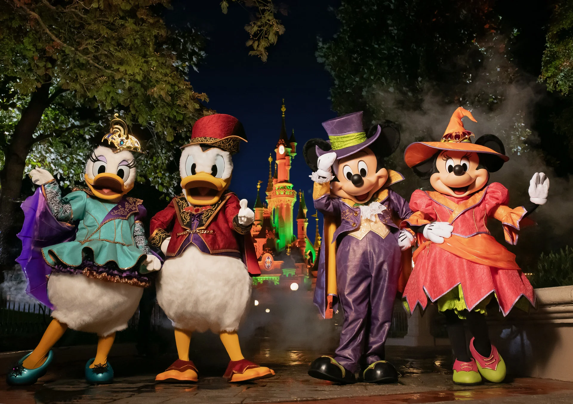 Quatre mascottes représentant des personnages Disney, vêtues de costumes d'Halloween, se tiennent devant un château illuminé de nuit, entourées d'arbres et de brouillard, incarnant ainsi l'atmosphère envoûtante du Festival d'Halloween à Disneyland Paris en 2024.