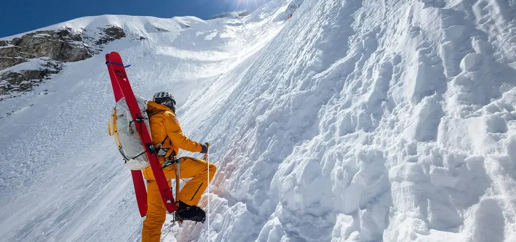 Un alpiniste vêtu d'une tenue orange gravit une pente enneigée et escarpée, ses skis rouges attachés à son sac à dos, dans une scène qui rappelle un documentaire du National Geographic, sous un soleil radieux et un ciel d'un bleu limpide.