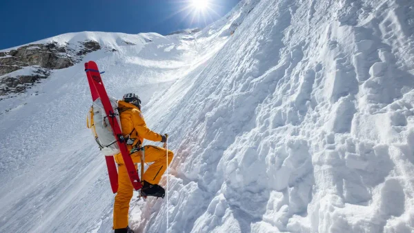 Un alpiniste vêtu d'une tenue orange gravit une pente enneigée et escarpée, ses skis rouges attachés à son sac à dos, dans une scène qui rappelle un documentaire du National Geographic, sous un soleil radieux et un ciel d'un bleu limpide.