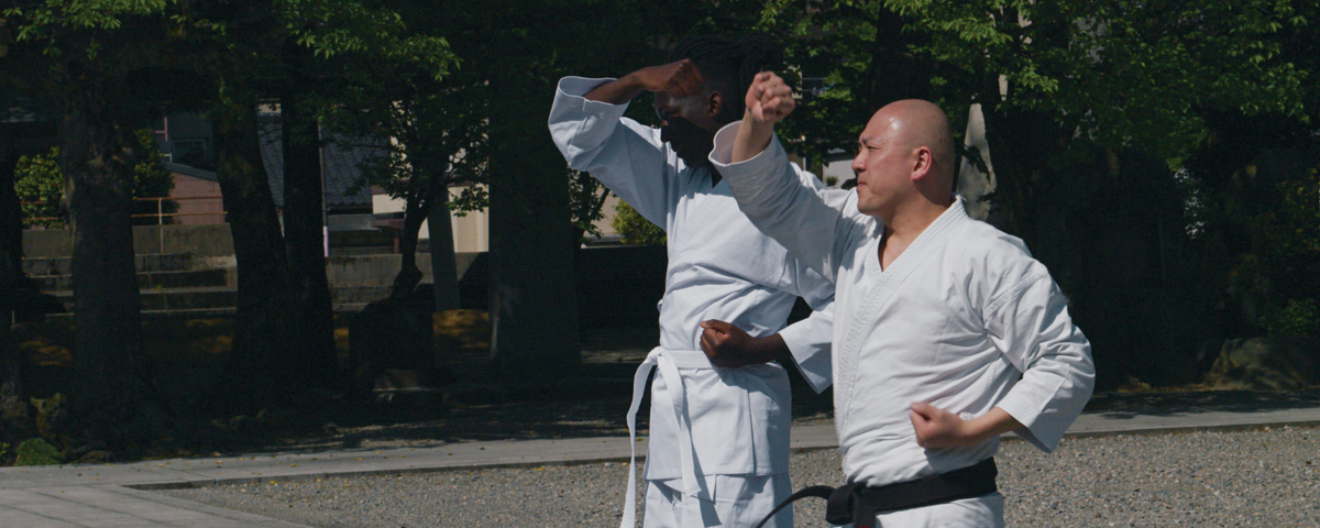 Deux personnes vêtues de kimonos de karaté blancs s'entraînent aux arts martiaux en plein air, exécutant des mouvements synchronisés, les bras levés et pliés, au milieu des arbres et d'une aire de gravier — une scène inspirante qui évoque la grâce disciplinée souvent représentée dans les films Disney+ ou mise à l'honneur dans les explorations de Mory Sacko sur la gastronomie japonaise.