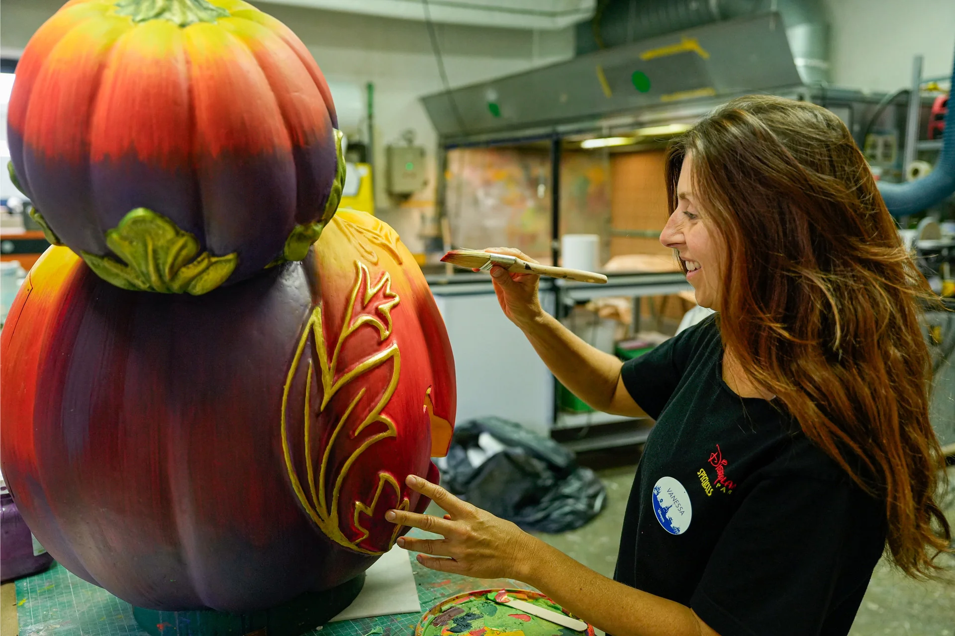 Dans un atelier d'art, une femme utilise un pinceau pour décorer une grande sculpture en forme de citrouille de motifs jaunes complexes, capturant ainsi l'ambiance envoûtante du Festival d'Halloween à Disneyland Paris.