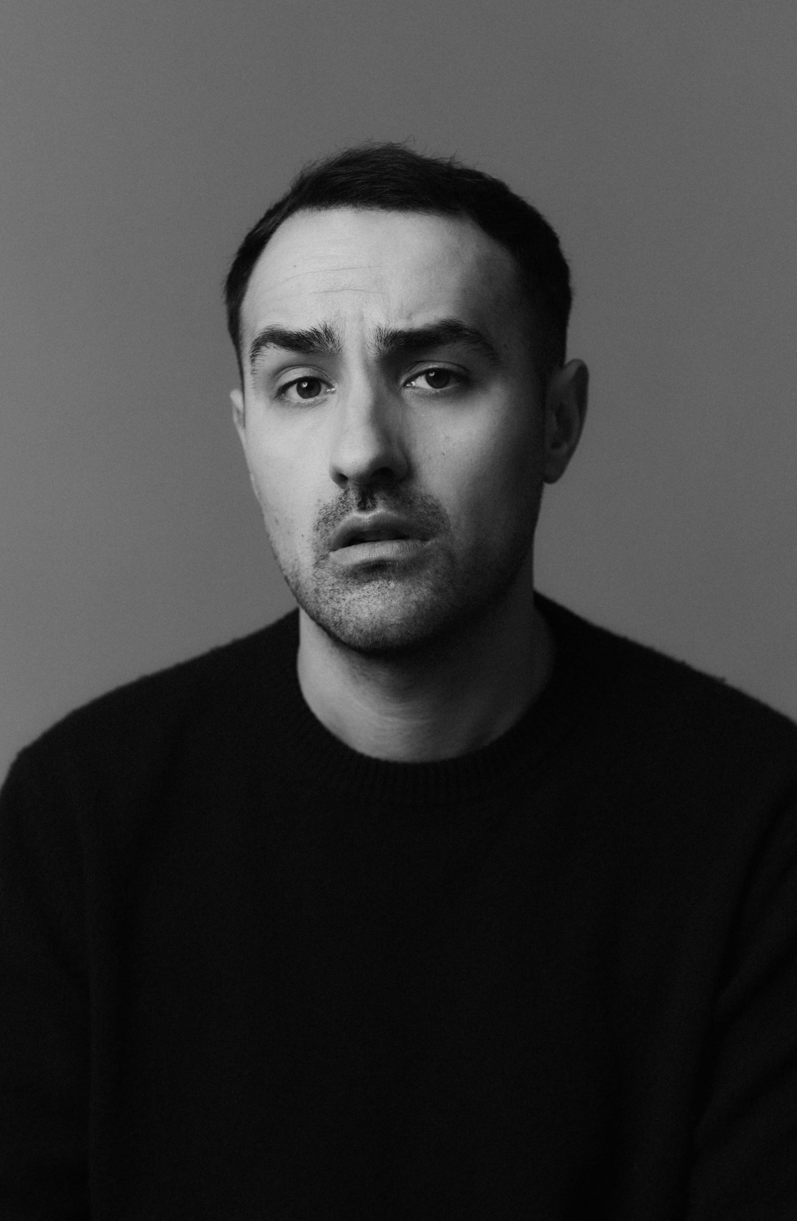 Portrait en noir et blanc d'un homme aux cheveux courts et foncés, arborant une moustache et une barbe soigneusement taillées, vêtu d'un pull à col rond foncé, l'air légèrement préoccupé devant un fond uni — une ambiance qui rappelle Brendan Gleeson dans l'un de ses rôles les plus contemplatifs.