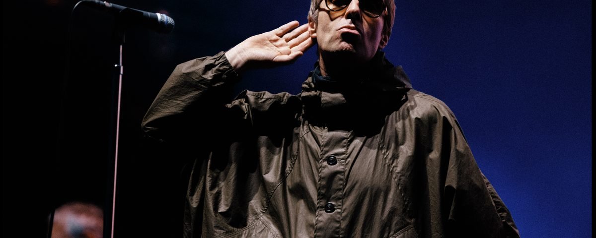 Une personne portant des lunettes de soleil et une veste sombre se tient sur scène, la main près de l'oreille, près d'un micro, avec un fond bleu derrière elle — rappelant le style emblématique d'Oasis interprétant « Don’t Look Back In Anger », comme on peut le voir dans les performances exclusives diffusées en streaming sur Disney+.