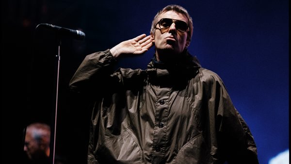 Une personne portant des lunettes de soleil et une veste sombre se tient sur scène, la main près de l'oreille, près d'un micro, avec un fond bleu derrière elle — rappelant le style emblématique d'Oasis interprétant « Don’t Look Back In Anger », comme on peut le voir dans les performances exclusives diffusées en streaming sur Disney+.