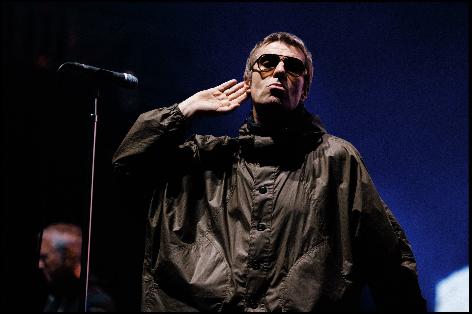 Une personne portant des lunettes de soleil et une veste sombre se tient sur scène, la main près de l'oreille, près d'un micro, avec un fond bleu derrière elle — rappelant le style emblématique d'Oasis interprétant « Don’t Look Back In Anger », comme on peut le voir dans les performances exclusives diffusées en streaming sur Disney+.