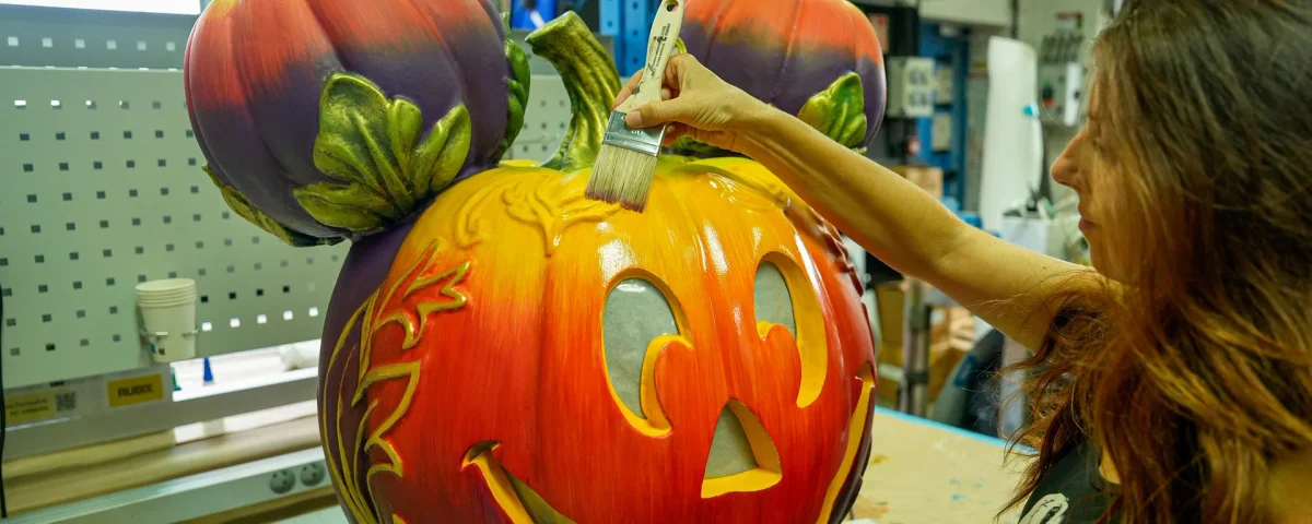 Dans un atelier, une femme peint les détails d'une grande sculpture en citrouille, dont les deux petites citrouilles servant d'oreilles font penser à une citrouille-lanterne à l'effigie de Mickey Mouse, incarnant ainsi l'esprit festif d'Halloween à Disneyland Paris.