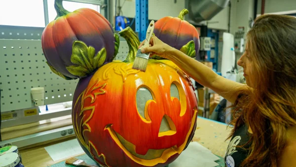 Dans un atelier, une femme peint les détails d'une grande sculpture en citrouille, dont les deux petites citrouilles servant d'oreilles font penser à une citrouille-lanterne à l'effigie de Mickey Mouse, incarnant ainsi l'esprit festif d'Halloween à Disneyland Paris.
