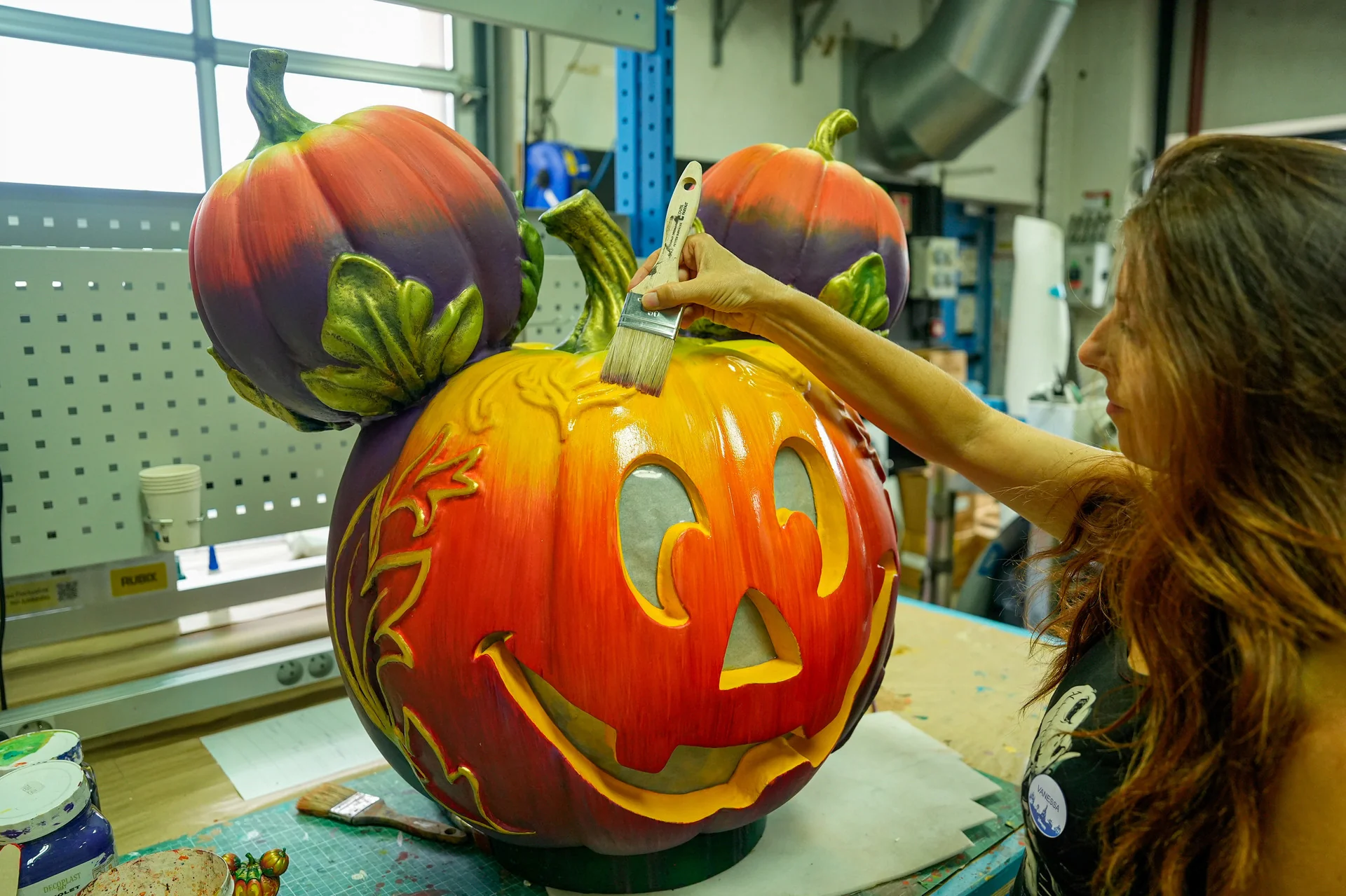 Dans un atelier, une femme peint les détails d'une grande sculpture en citrouille, dont les deux petites citrouilles servant d'oreilles font penser à une citrouille-lanterne à l'effigie de Mickey Mouse, incarnant ainsi l'esprit festif d'Halloween à Disneyland Paris.