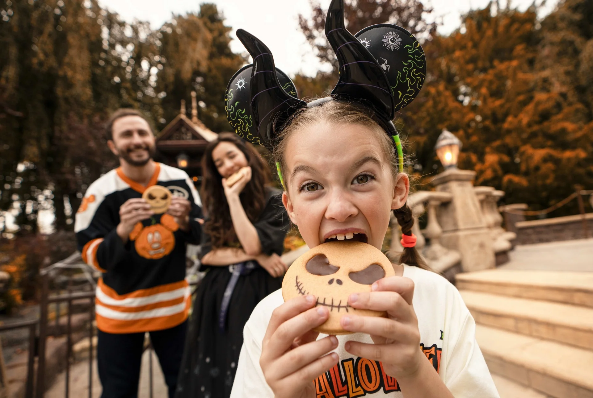 Une fillette déguisée pour Halloween croque dans un biscuit à l'effigie de Jack Skellington, illustrant ainsi l'ambiance festive du Festival Halloween à Disneyland Paris, tandis qu'à l'arrière-plan, flou et en extérieur, deux adultes souriants tiennent des biscuits à la main.