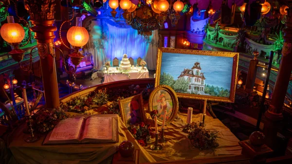 Une table décorée de tableaux encadrés, de bougies et d'un livre ouvert donne sur une scène aménagée pour un événement solennel, baignée d'une lumière chaleureuse et colorée qui évoque l'ambiance enchanteresse qui fait la renommée d'Halloween à Disneyland Paris.
