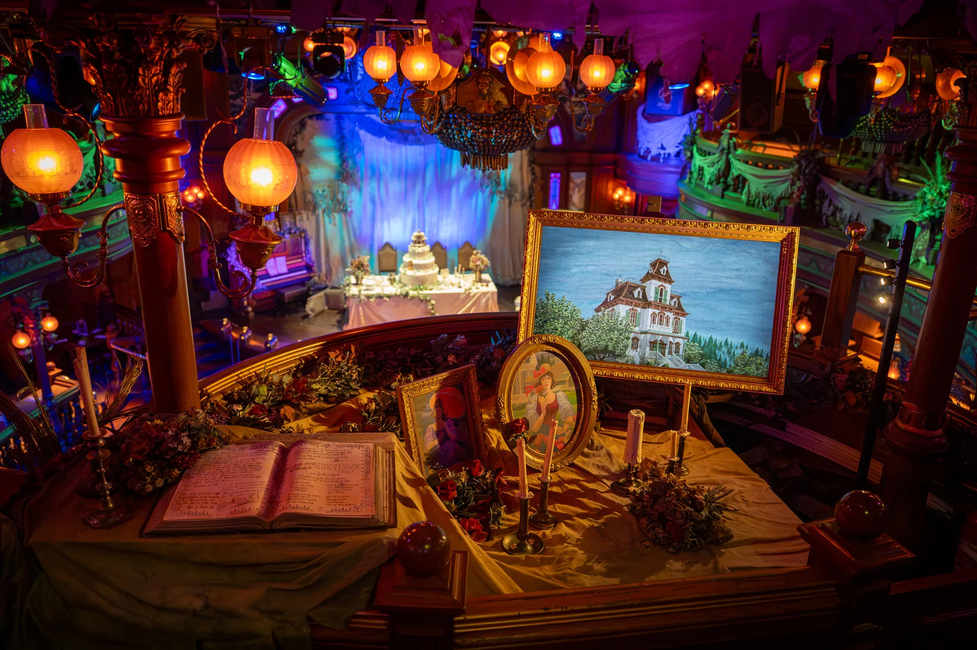 Une table décorée de tableaux encadrés, de bougies et d'un livre ouvert donne sur une scène aménagée pour un événement solennel, baignée d'une lumière chaleureuse et colorée qui évoque l'ambiance enchanteresse qui fait la renommée d'Halloween à Disneyland Paris.