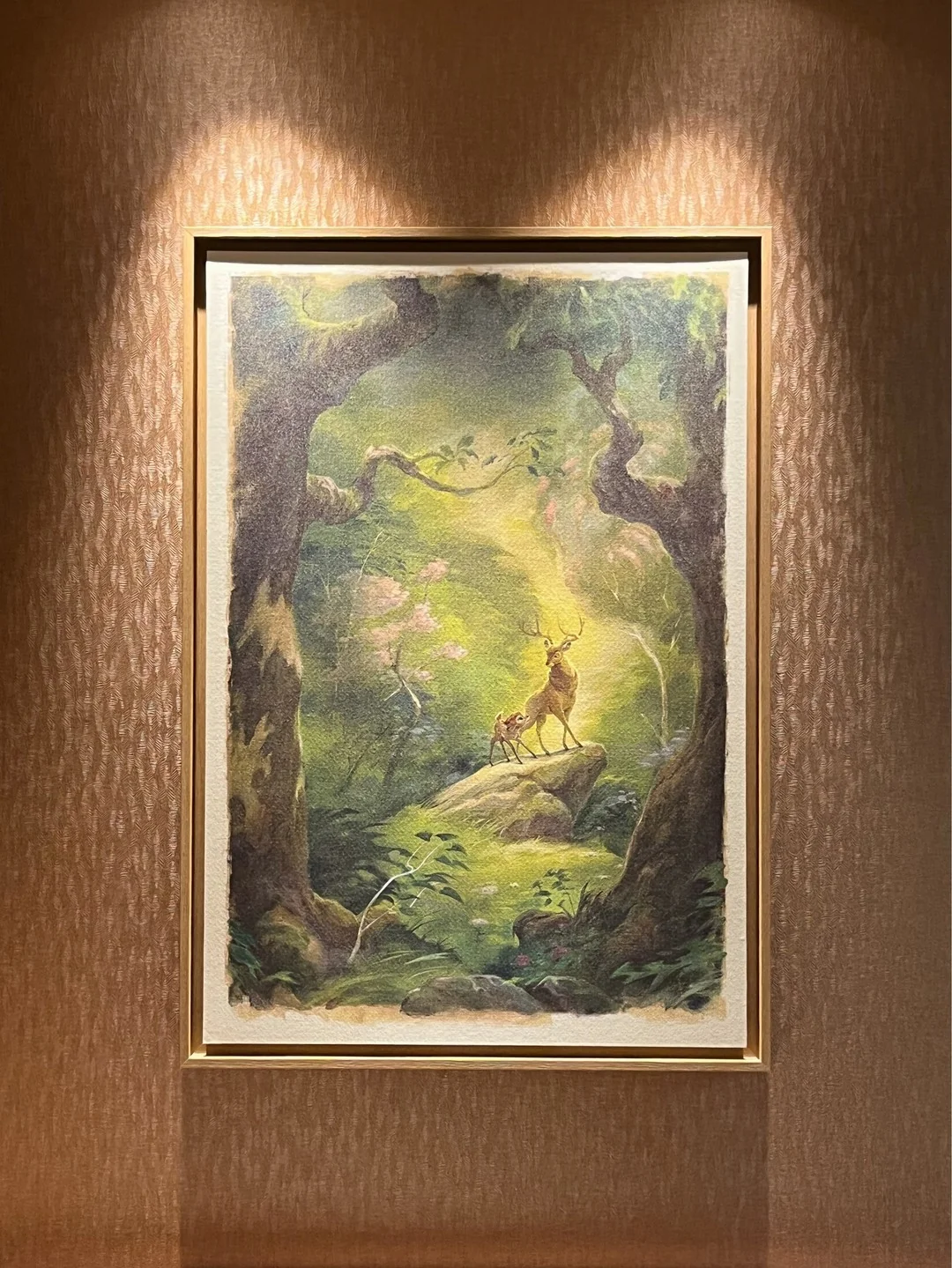 Tableau encadré représentant un cerf et son faon debout sur un rocher dans une forêt baignée de soleil, qui rappelle les « tableaux exclusifs » que l'on trouve dans les « chambres rénovées » du Disney Sequoia Lodge, accroché sur un mur à motifs sous des spots.