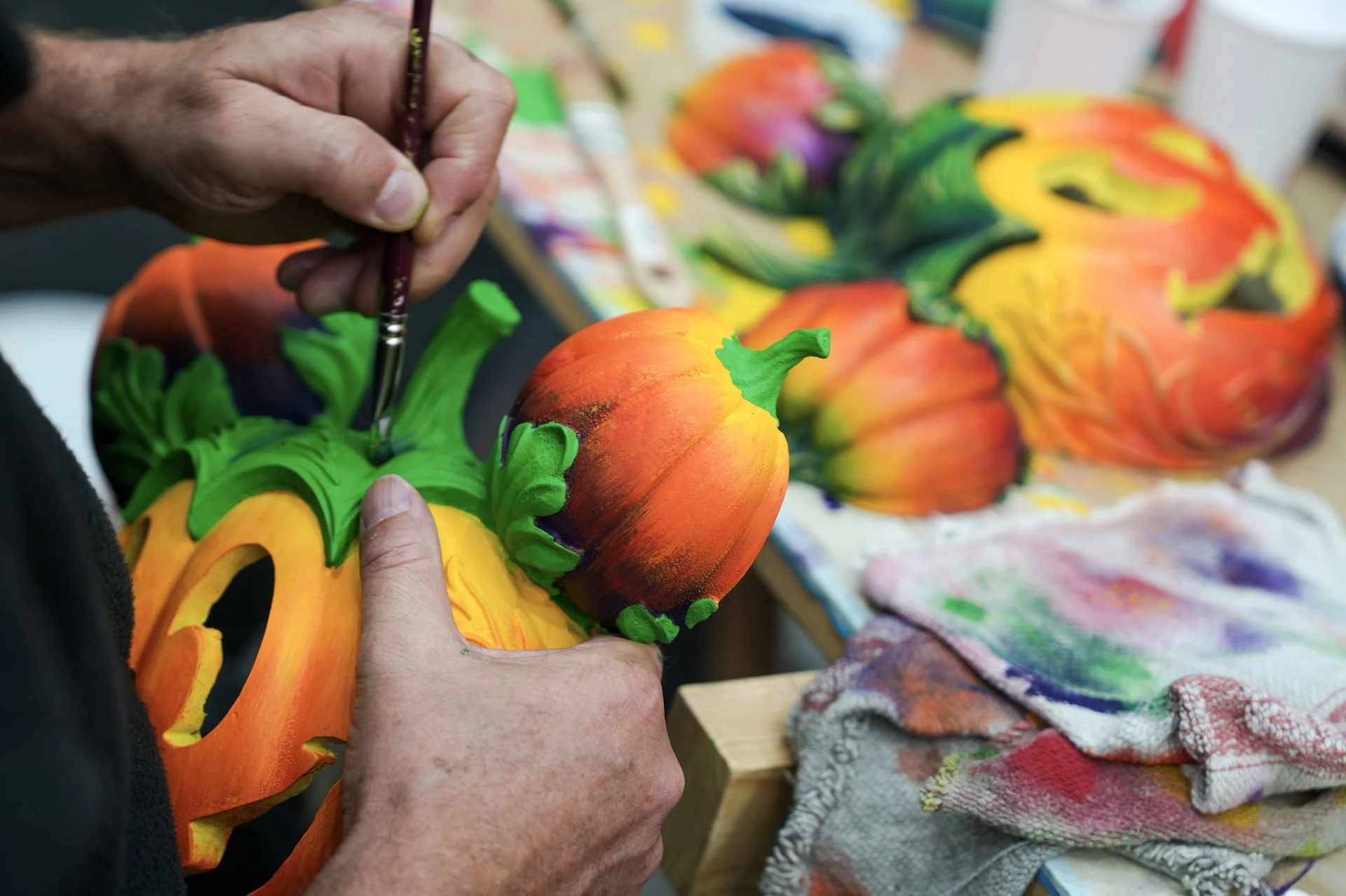 Une personne utilise un pinceau pour appliquer de la peinture verte sur la tige d'une citrouille décorative, inspirée du Festival Halloween Disney, à côté d'autres citrouilles décorées et d'un tissu coloré.