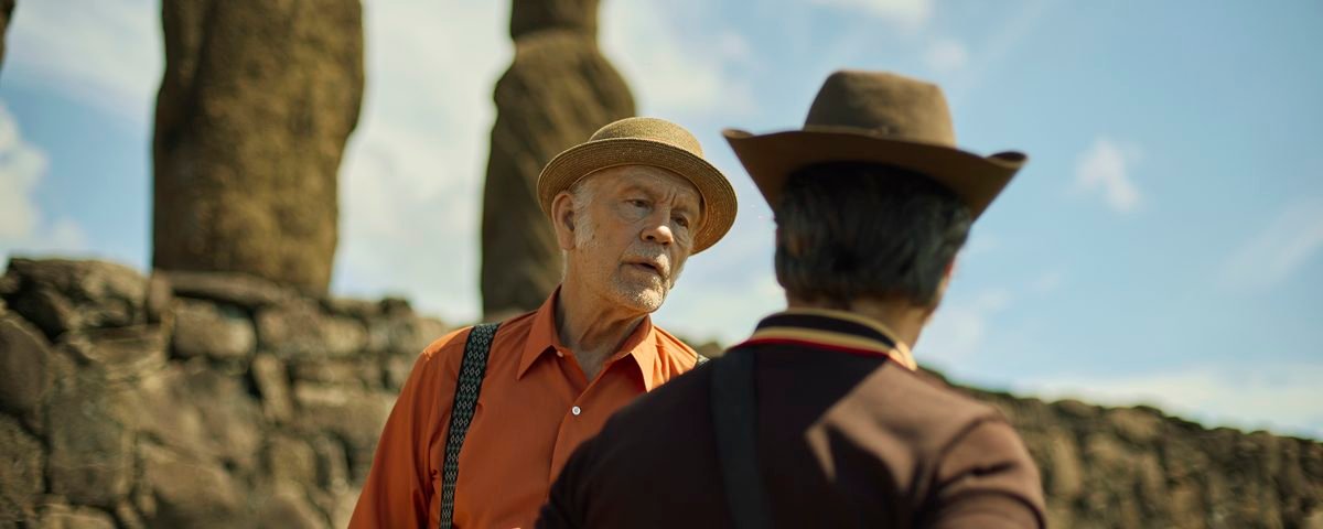 Deux hommes coiffés de chapeaux discutent devant de grandes statues de pierre, sur un site archéologique en plein air, sous un ciel bleu parsemé de nuages, dans une scène qui rappelle un film de Martin McDonagh.