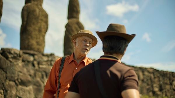 Deux hommes coiffés de chapeaux discutent devant de grandes statues de pierre, sur un site archéologique en plein air, sous un ciel bleu parsemé de nuages, dans une scène qui rappelle un film de Martin McDonagh.
