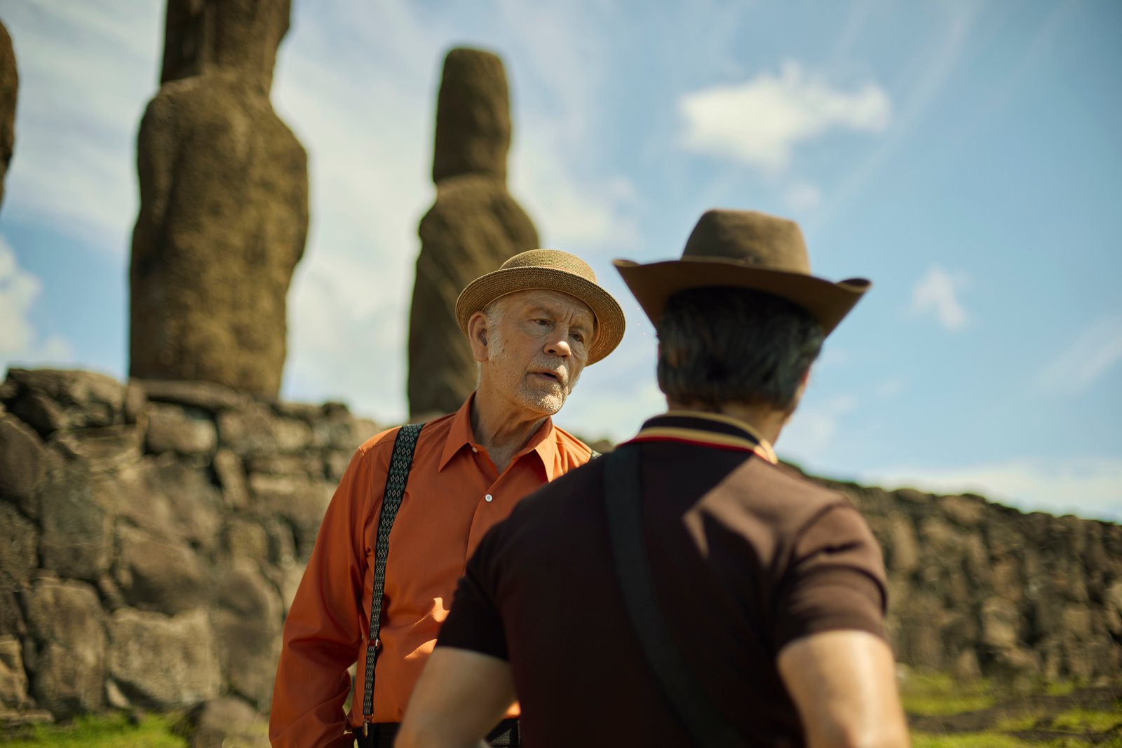 Deux hommes coiffés de chapeaux discutent devant de grandes statues de pierre, sur un site archéologique en plein air, sous un ciel bleu parsemé de nuages, dans une scène qui rappelle un film de Martin McDonagh.