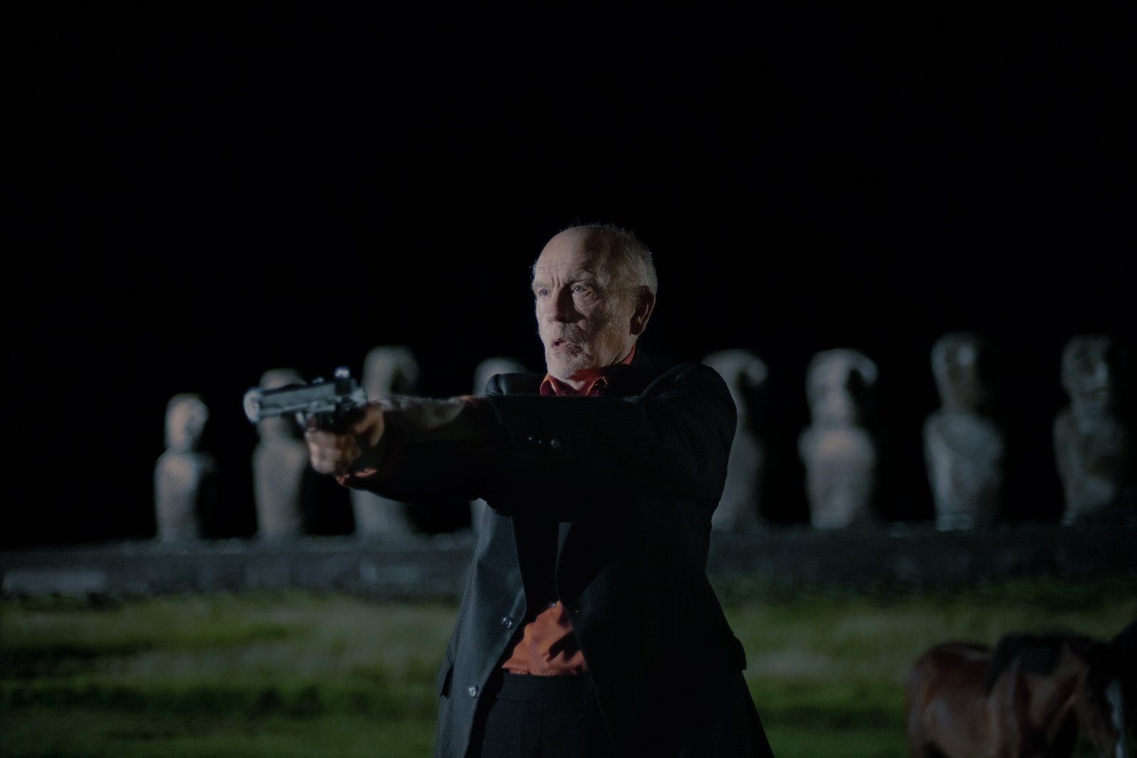 Un homme d’un certain âge en costume, qui ressemble à John Malkovich, pointe un pistolet vers l’avant, de nuit, debout à l’extérieur, avec de grandes statues de pierre en arrière-plan — une image qui évoque une scène tendue tirée d’un thriller de Martin McDonagh ou peut-être de *Wild Horse Nine*.