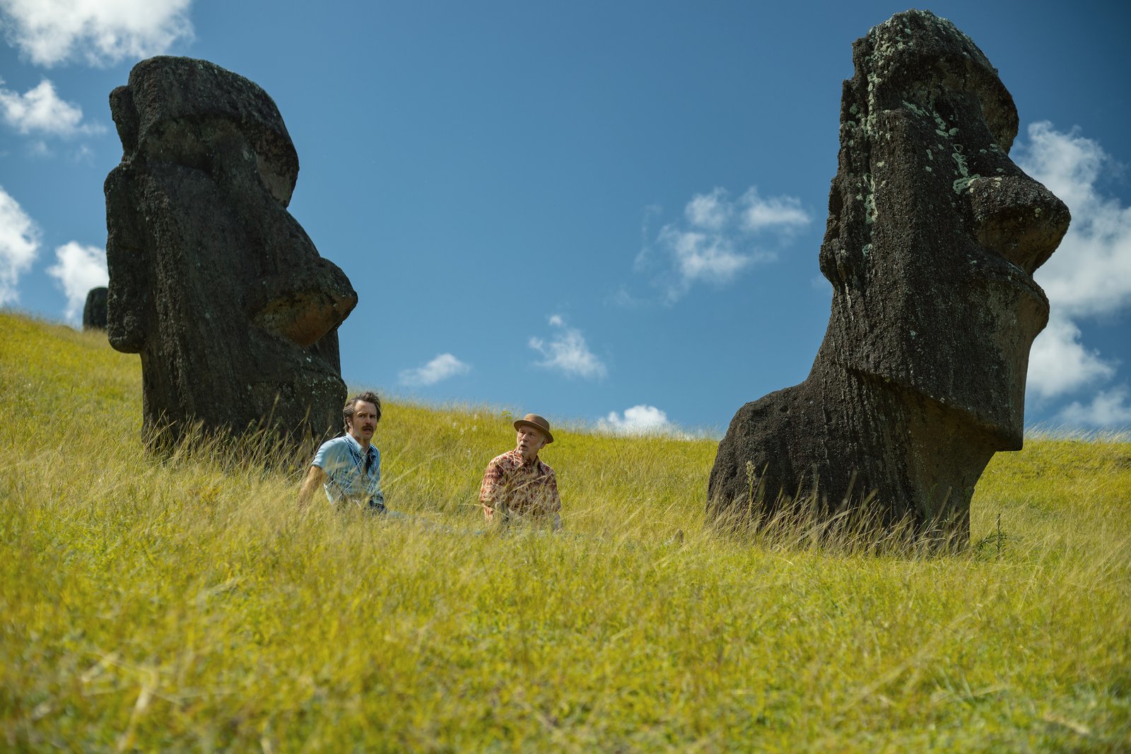 Deux personnes sont assises dans les hautes herbes, entre deux grandes statues de moai en pierre, sur un versant ensoleillé, sous un ciel bleu parsemé de nuages, créant une scène paisible qui rappelle les paysages cinématographiques de Martin McDonagh.