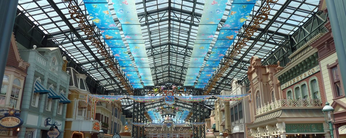 La foule se promène dans une galerie marchande couverte dont le plafond de verre est orné de panneaux bleus et de motifs colorés, entourée de vitrines aux façades thématiques et richement décorées qui rappellent Tokyo Disneyland, et bordée de boutiques de souvenirs, pour une expérience de shopping unique.