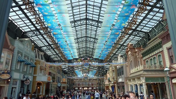La foule se promène dans une galerie marchande couverte dont le plafond de verre est orné de panneaux bleus et de motifs colorés, entourée de vitrines aux façades thématiques et richement décorées qui rappellent Tokyo Disneyland, et bordée de boutiques de souvenirs, pour une expérience de shopping unique.
