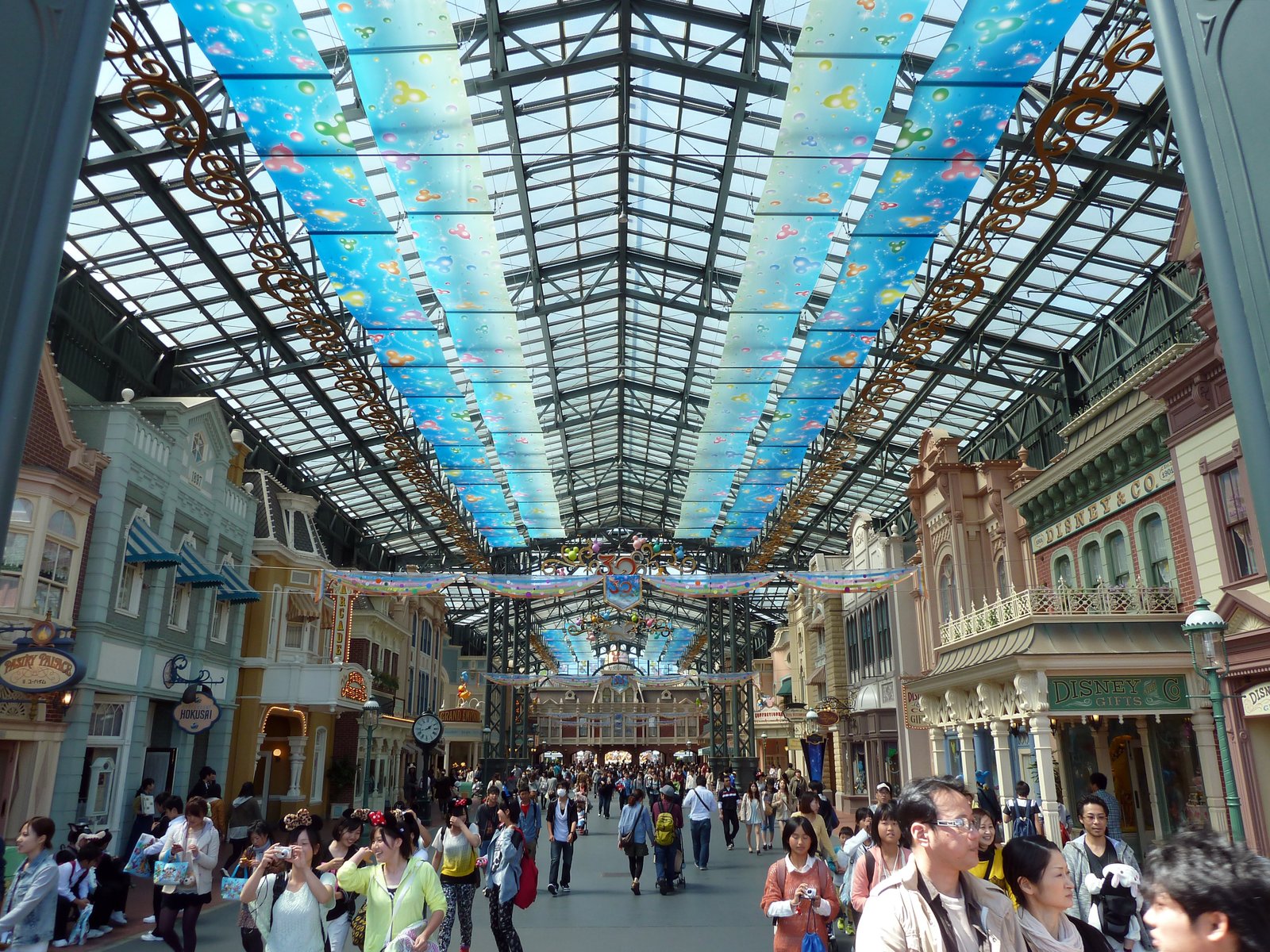 La foule se promène dans une galerie marchande couverte dont le plafond de verre est orné de panneaux bleus et de motifs colorés, entourée de vitrines aux façades thématiques et richement décorées qui rappellent Tokyo Disneyland, et bordée de boutiques de souvenirs, pour une expérience de shopping unique.
