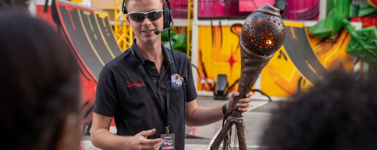 Une personne portant des lunettes de soleil et un casque audio s'adresse à un groupe tout en tenant un grand bâton sculpté, dans un espace extérieur orné de fresques colorées et de rampes. Elle coordonne le travail des équipes d'effets spéciaux afin de créer des effets éblouissants qui rappellent ceux que l'on peut voir à Disneyland Paris.