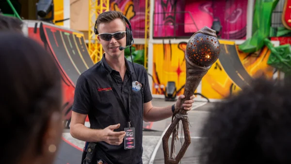 Une personne portant des lunettes de soleil et un casque audio s'adresse à un groupe tout en tenant un grand bâton sculpté, dans un espace extérieur orné de fresques colorées et de rampes. Elle coordonne le travail des équipes d'effets spéciaux afin de créer des effets éblouissants qui rappellent ceux que l'on peut voir à Disneyland Paris.