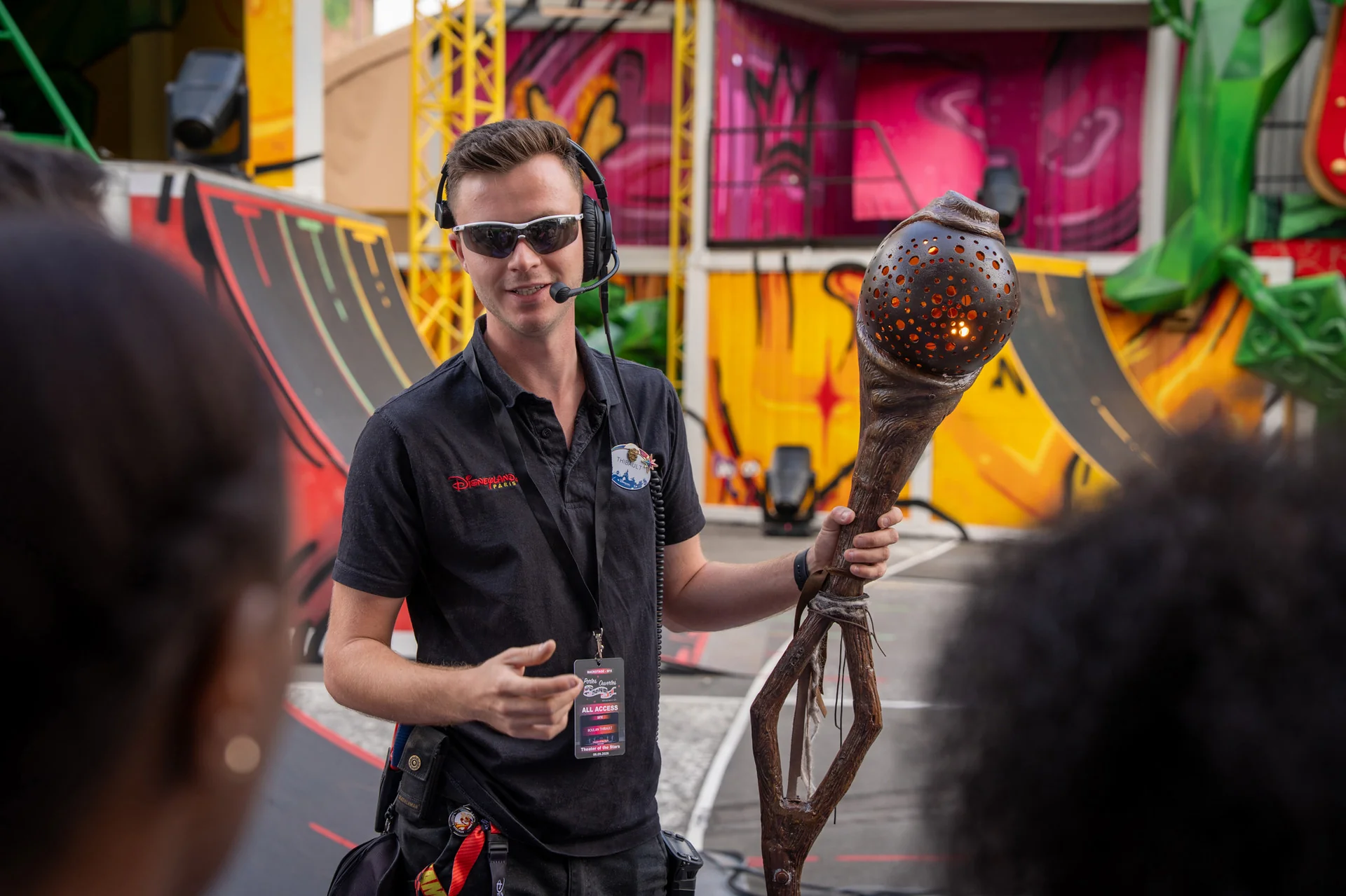 Une personne portant des lunettes de soleil et un casque audio s'adresse à un groupe tout en tenant un grand bâton sculpté, dans un espace extérieur orné de fresques colorées et de rampes. Elle coordonne le travail des équipes d'effets spéciaux afin de créer des effets éblouissants qui rappellent ceux que l'on peut voir à Disneyland Paris.