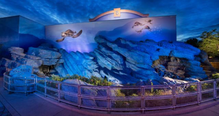 Extérieur rocheux et baigné d'une lumière bleue d'une exposition d'aquarium ornée de deux fresques représentant des tortues sur le mur — un hommage à « Crush’s Coaster » —, entourée d'une balustrade et de plantes sous un ciel crépusculaire, évoquant l'effervescence de son inauguration le 9 juin 2007.