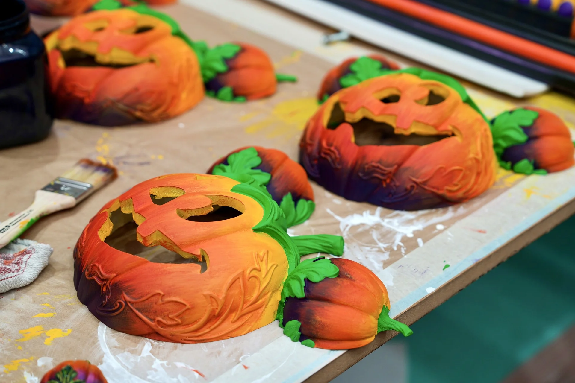 Trois masques de citrouille peints, aux visages sculptés et ornés de feuilles vertes, reposent sur une table de travail aux côtés d'un pinceau et de matériel artistique, créant ainsi une ambiance d'Halloween envoûtante qui rappelle le Festival d'Halloween de Disney à Disneyland Paris.