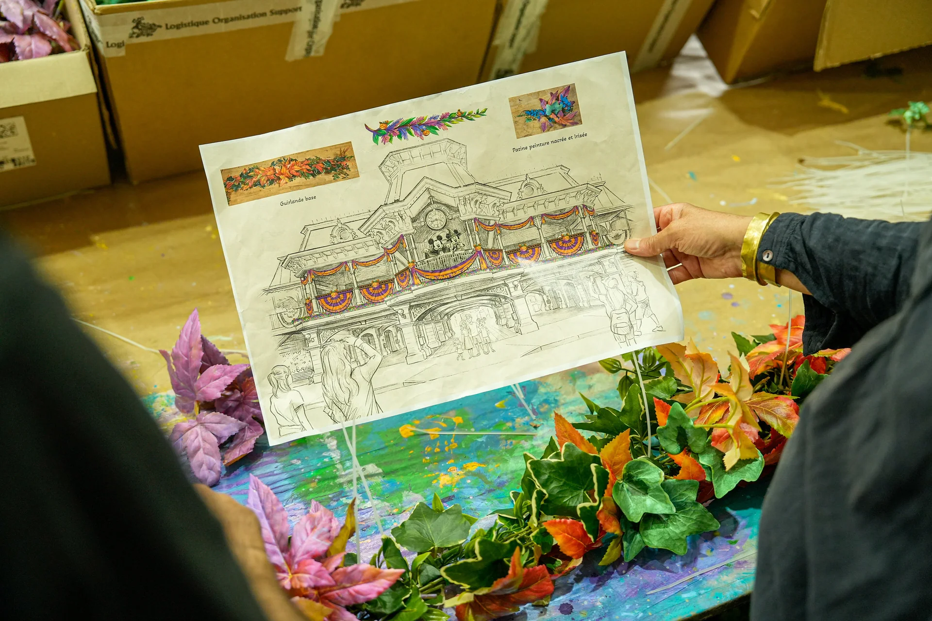 Une personne tient un dessin représentant un bâtiment décoré, entouré de feuilles d'automne artificielles et de boîtes en arrière-plan, illustrant l'ambiance enchanteresse d'Halloween Disney telle qu'on peut la découvrir à Disneyland Paris pendant la période des fêtes.