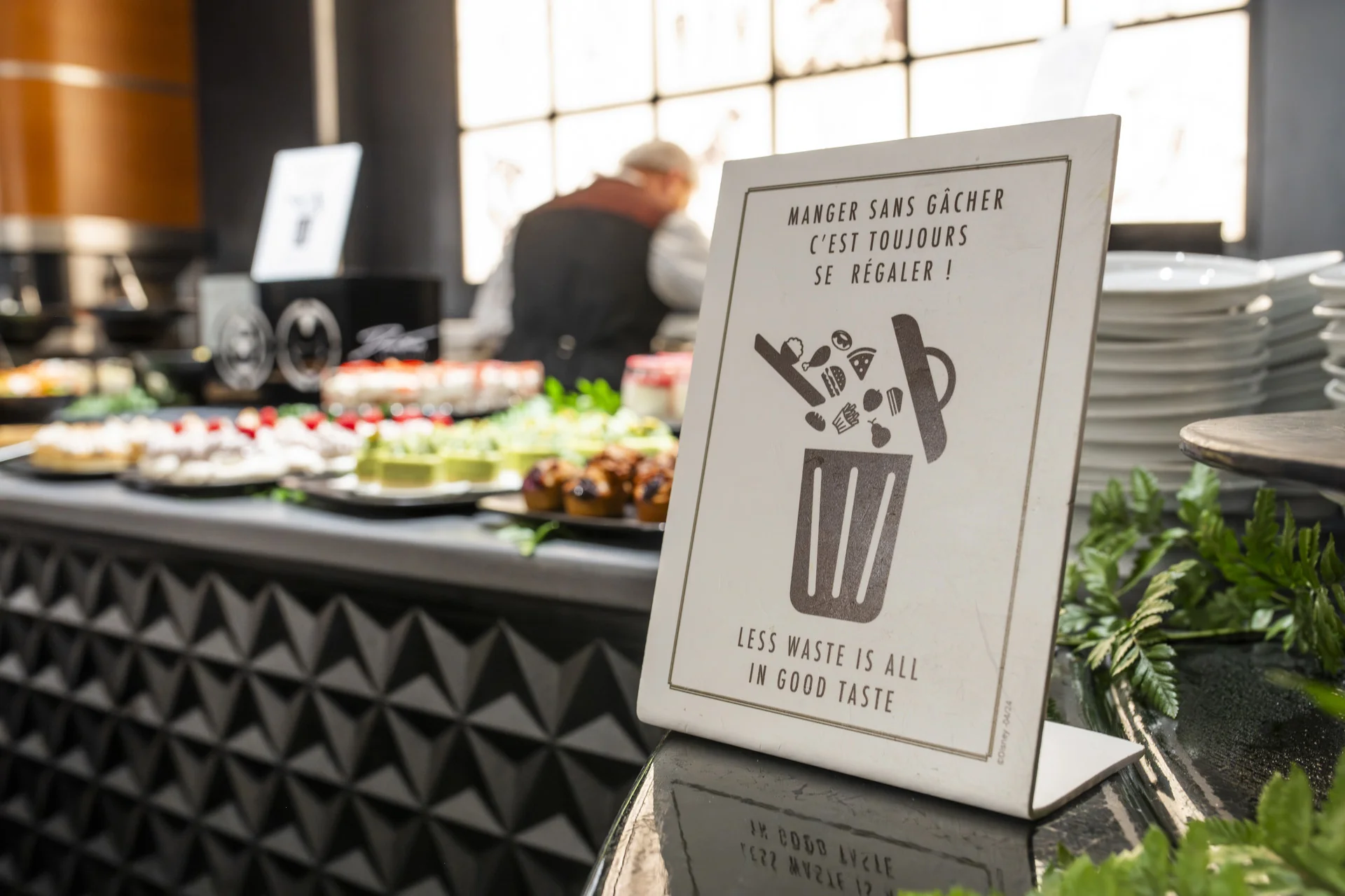 Une affiche placée près d’un buffet de Disneyland Paris, réalisée par Morgane Pelletier, porte l’inscription « Moins de gaspillage, c’est une question de bon goût », illustrant ainsi l’engagement environnemental du restaurant. On y voit des assiettes, des desserts et un client.