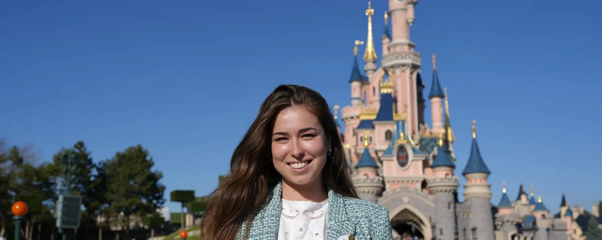 Morgane Pelletier, vêtue d'un blazer léger, pose en souriant devant le château de conte de fées rose de Disneyland Paris, par une journée ensoleillée.