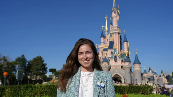 Morgane Pelletier, vêtue d'un blazer léger, pose en souriant devant le château de conte de fées rose de Disneyland Paris, par une journée ensoleillée.