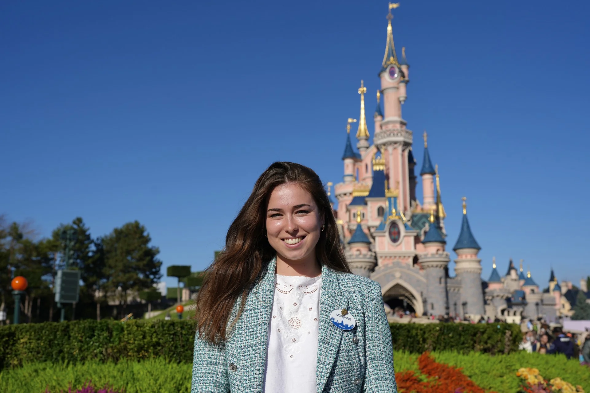 Morgane Pelletier, vêtue d'un blazer léger, pose en souriant devant le château de conte de fées rose de Disneyland Paris, par une journée ensoleillée.