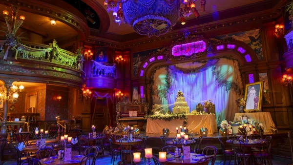 Sommier et faiblement éclairé, le saloon « Unlucky Nugget » se métamorphose pour Halloween 2026, avec des tables rondes, des bougies et des décorations soignées un peu partout. Une scène décorée met en valeur un grand gâteau et un portrait encadré, le tout dans un style vintage et théâtral rehaussé d’un éclairage coloré.