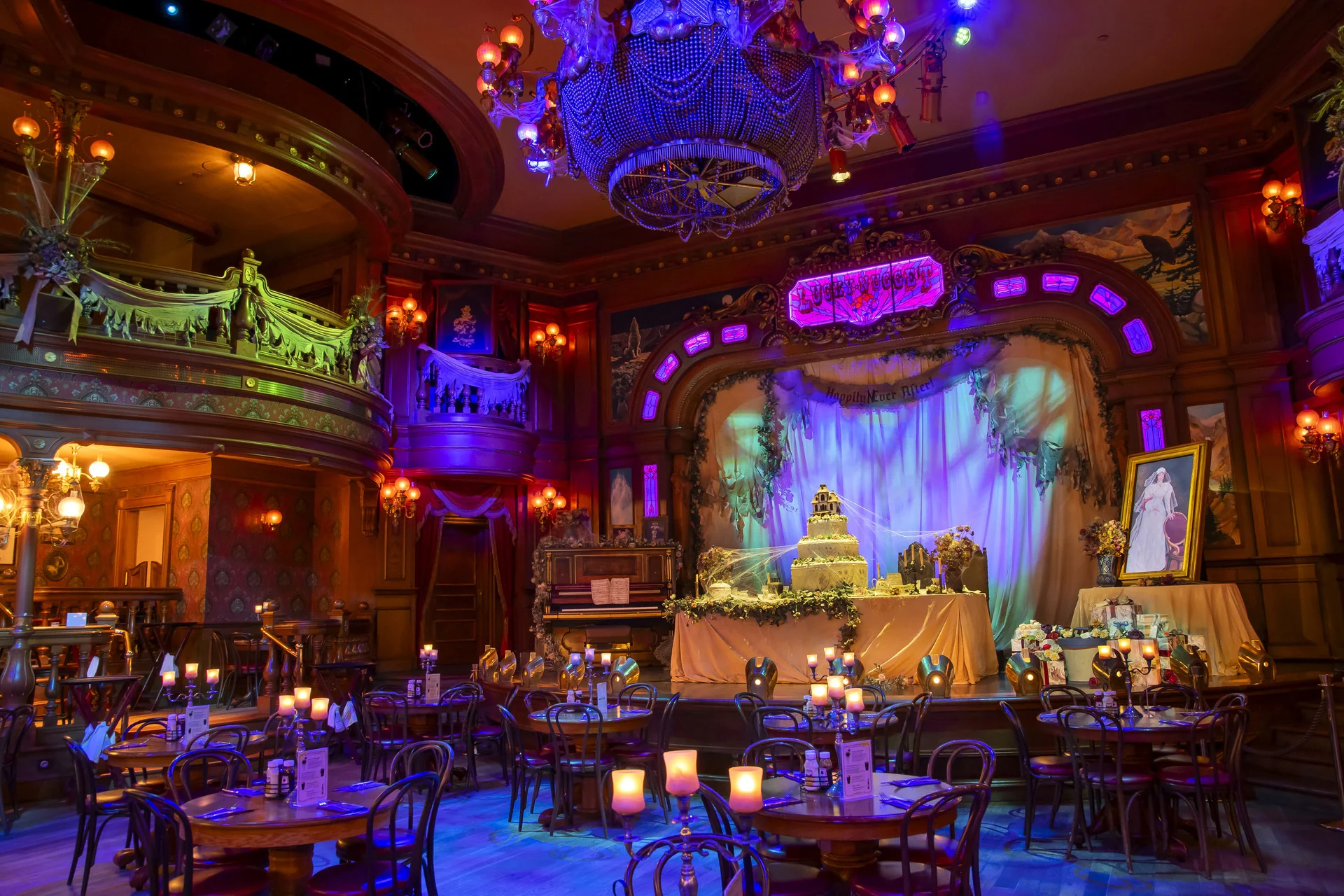 Sommier et faiblement éclairé, le saloon « Unlucky Nugget » se métamorphose pour Halloween 2026, avec des tables rondes, des bougies et des décorations soignées un peu partout. Une scène décorée met en valeur un grand gâteau et un portrait encadré, le tout dans un style vintage et théâtral rehaussé d’un éclairage coloré.