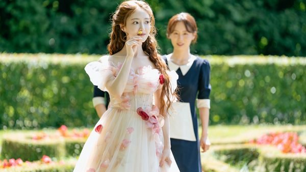 Une femme vêtue d'une robe à fleurs « L'Impératrice Remariée » porte une fleur à son nez, tandis qu'une femme en uniforme de femme de chambre se tient derrière elle dans un jardin.