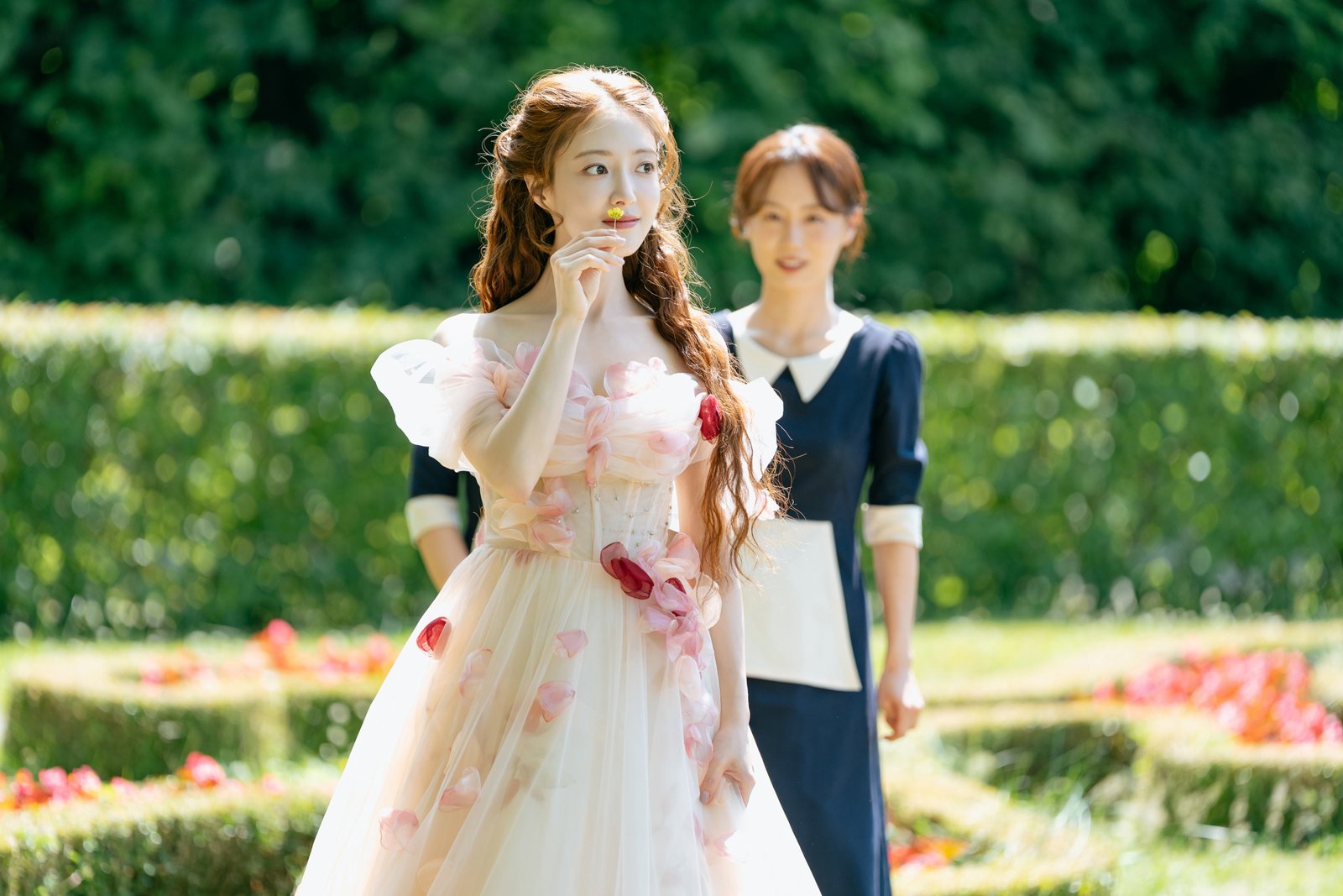 Une femme vêtue d'une robe à fleurs « L'Impératrice Remariée » porte une fleur à son nez, tandis qu'une femme en uniforme de femme de chambre se tient derrière elle dans un jardin.