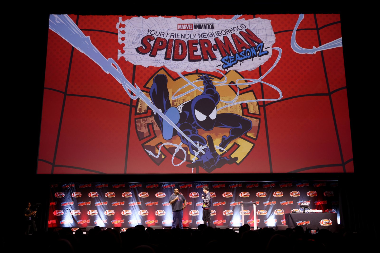 Deux personnes sur la scène du Comic Con, devant un grand écran affichant le logo de la saison 2 de la série « Your Friendly Neighborhood Spider-Man » de Marvel.