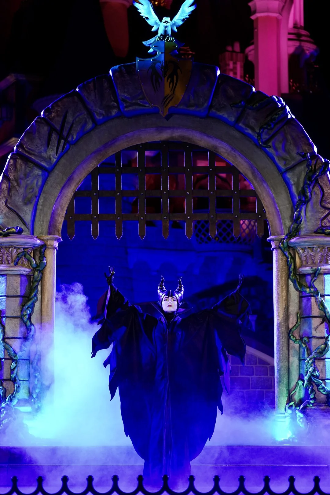 Maleficent, vêtue d'une robe noire et coiffée d'un casque à cornes, se tient les bras tendus sous une arche en pierre envahie par le brouillard d'Halloween 2026.