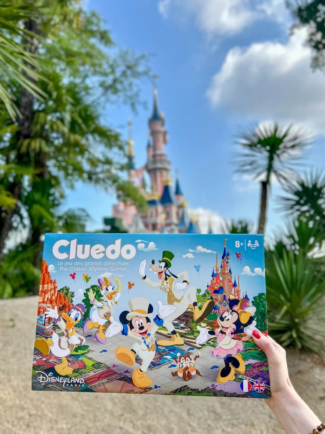 Une personne tient entre ses mains une édition exclusive du jeu de société « Cluedo » de Disney devant le château de la Belle au bois dormant à Disneyland Paris.