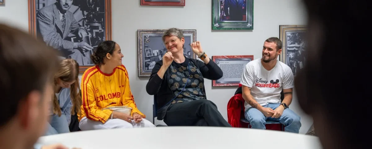 Un groupe de personnes est assis en cercle, discutant et souriant dans une pièce décorée de photos encadrées, dans une ambiance chaleureuse et conviviale.