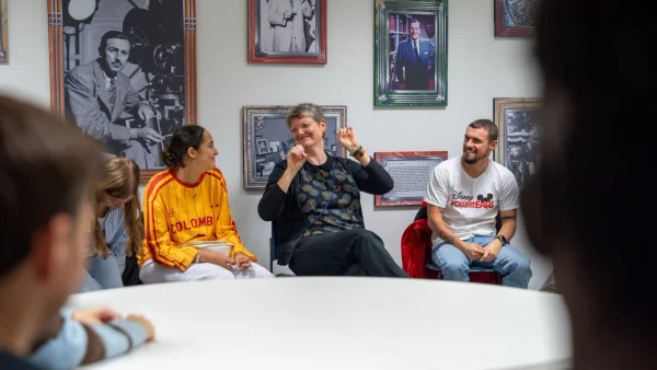Un groupe de personnes est assis en cercle, discutant et souriant dans une pièce décorée de photos encadrées, dans une ambiance chaleureuse et conviviale.