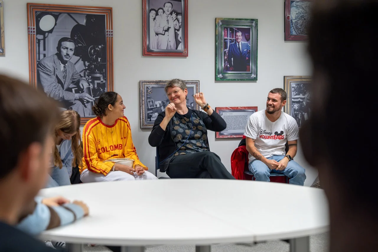Un groupe de personnes est assis en cercle, discutant et souriant dans une pièce décorée de photos encadrées, dans une ambiance chaleureuse et conviviale.
