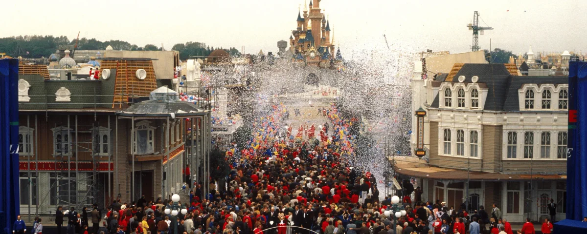 Une foule nombreuse envahit une rue de Disneyland Paris, tandis que des confettis tombent devant le Château de la Belle au Bois Dormant.